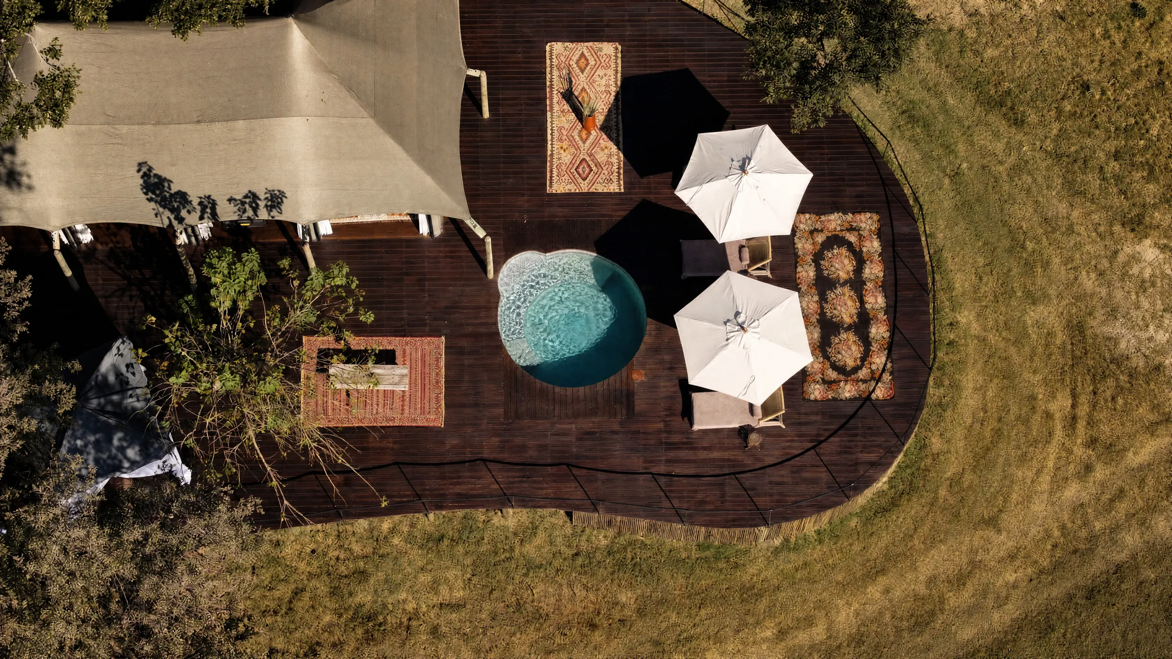 A villa pool deck seen from above, two white parasols, loungers and rugs on the dark wood, the grass all around