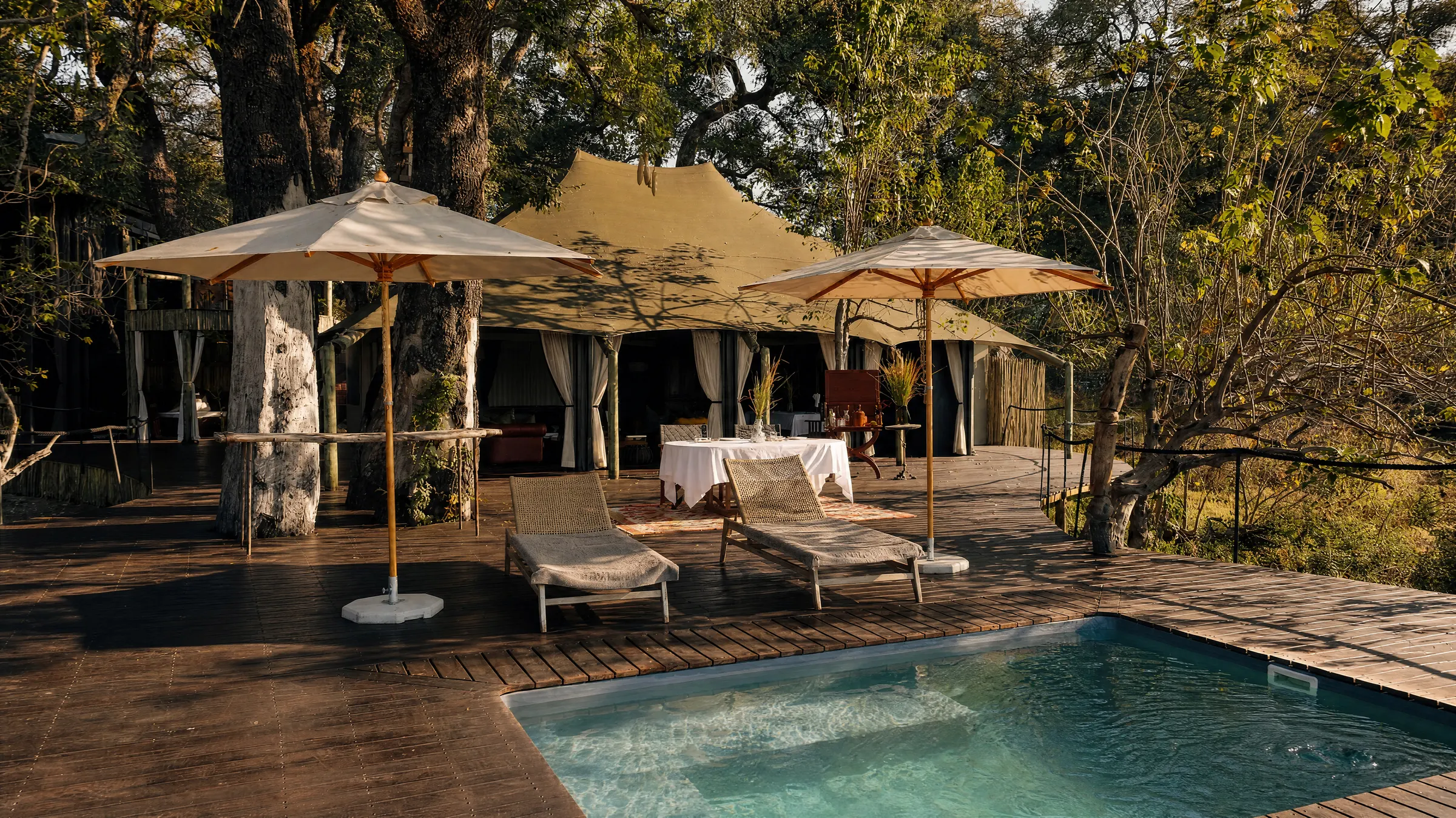 The pool of a villa in the sun, loungers and two parasols on the deck and a table laid under the canvas