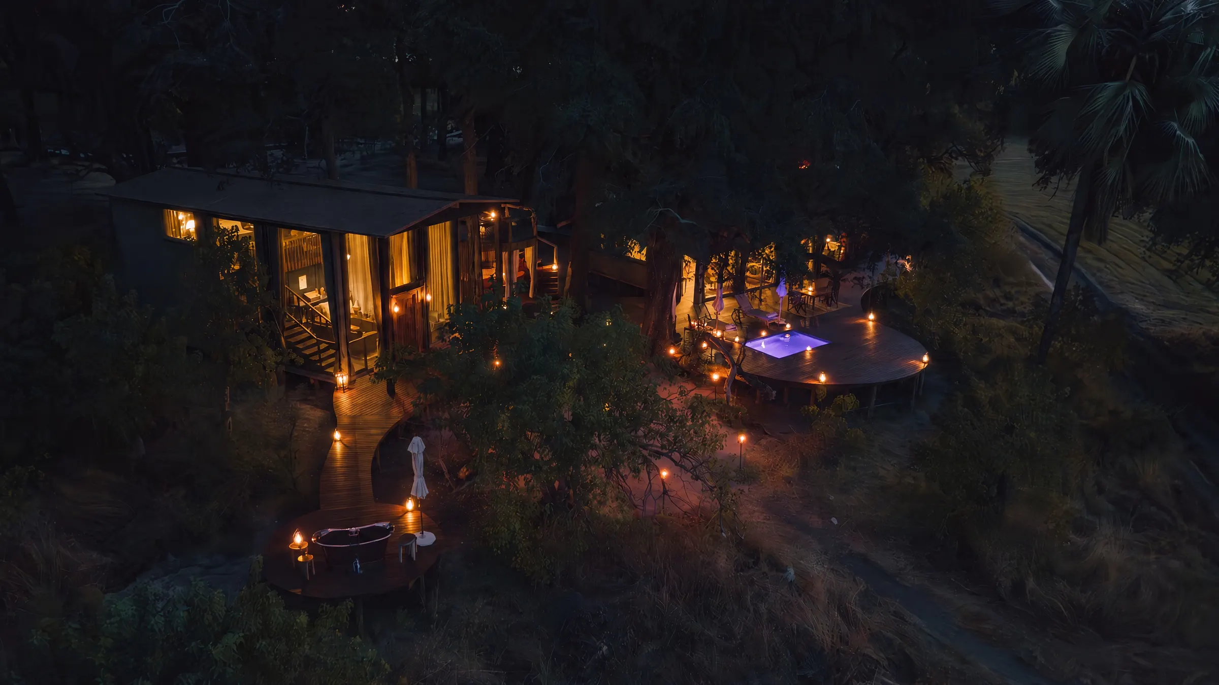 A villa seen from the air at nightfall, its windows lit, lanterns along the walkways and the pool glowing on the deck