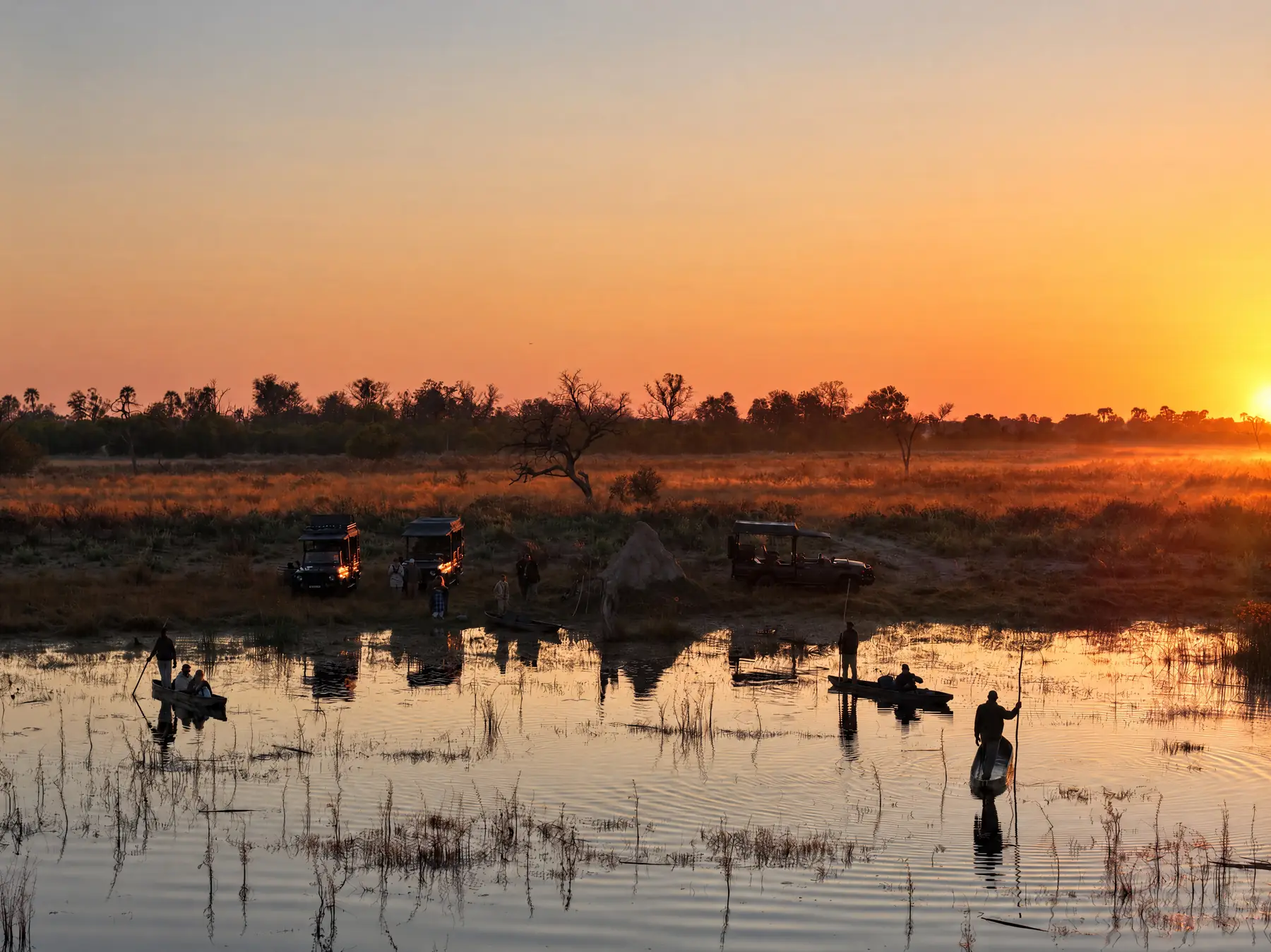 The sun setting over open water, with mokoros and their polers in the shallows and game vehicles on the bank
