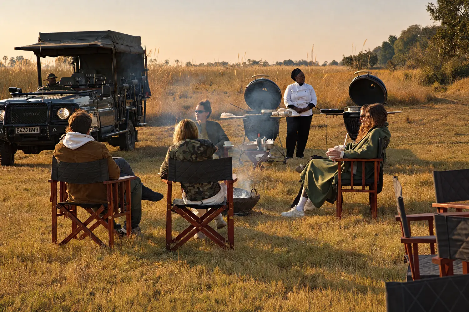 Guests in safari chairs around a fire on the plain, the trucks behind them