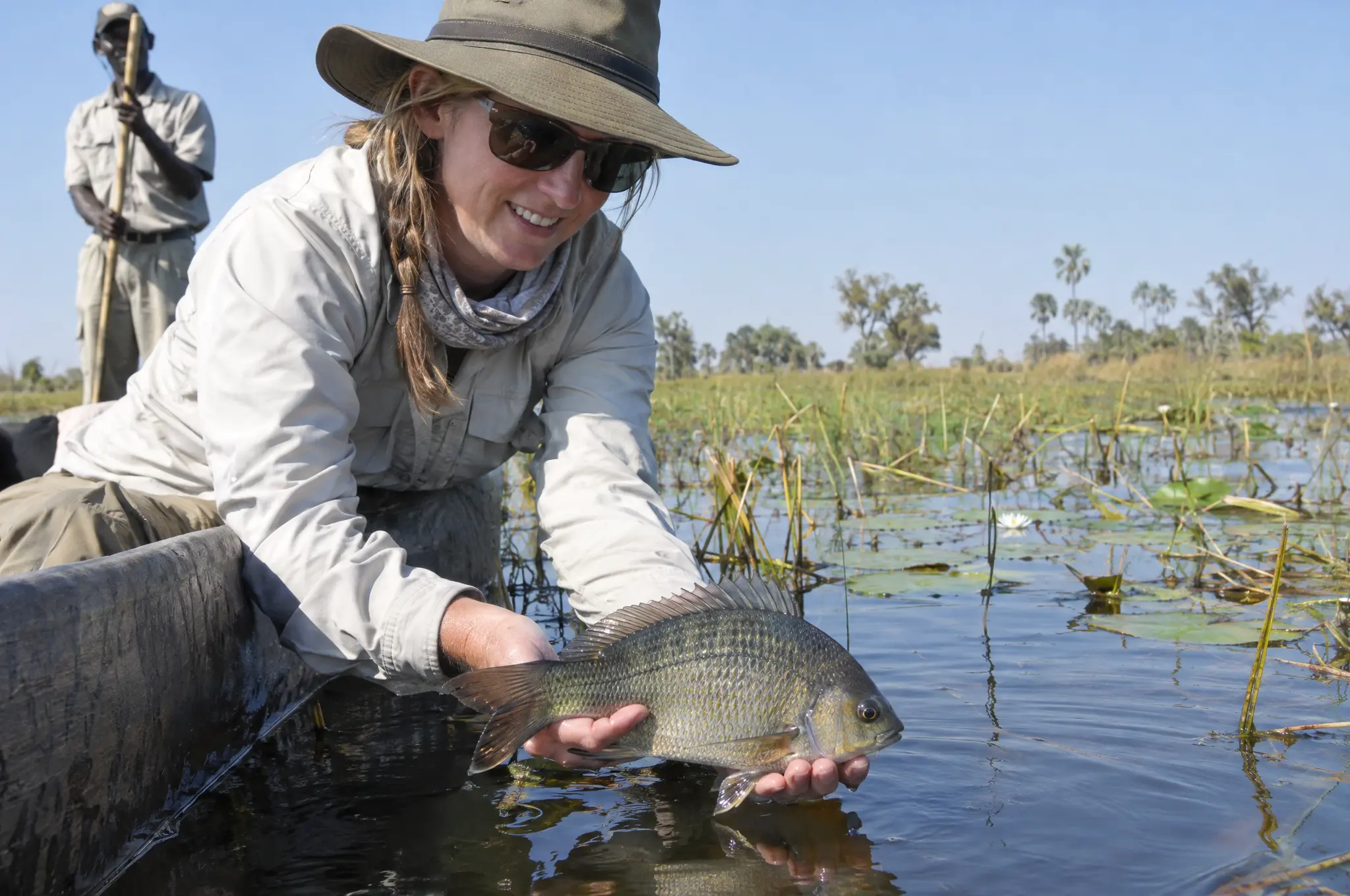 A smiling guest in a sun hat holding a bream over the side of a mokoro, the poler standing behind her