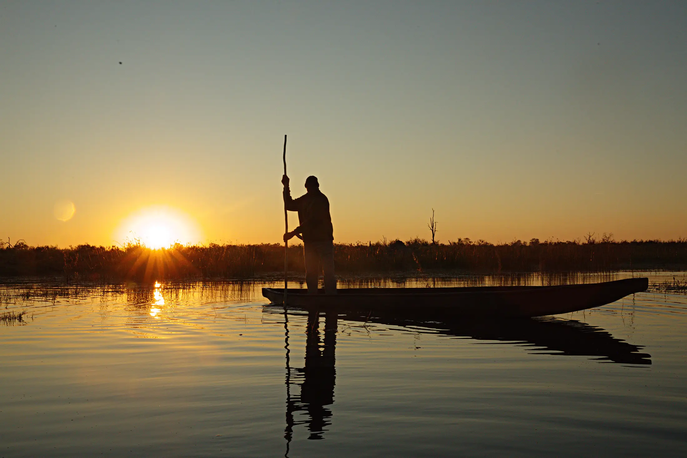 A poler standing in his mokoro against the rising sun, the water still around him