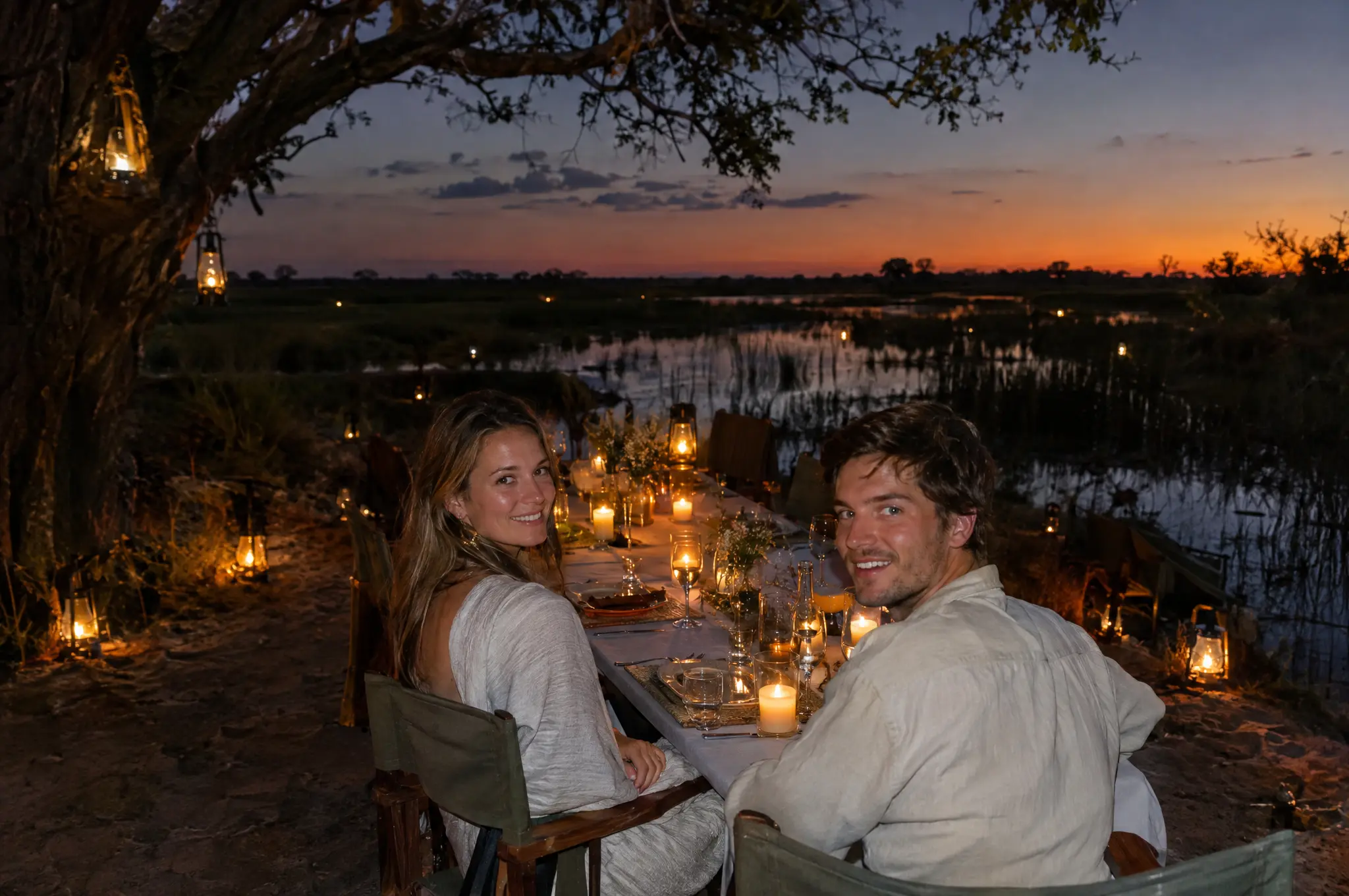 A couple smiling at a long candlelit table under a tree on a small island at dusk, lanterns all around and the water behind