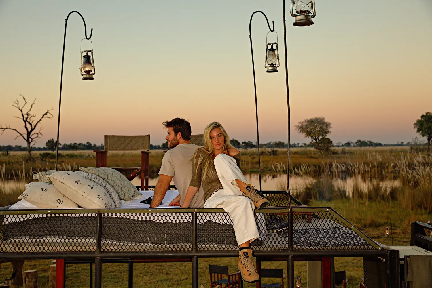 Two guests sitting on the sleep out bed at dusk, lanterns on their poles around them