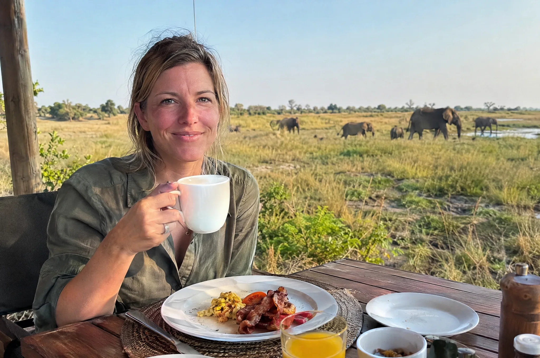 A guest smiling over her coffee at breakfast, elephants walking past in the distance