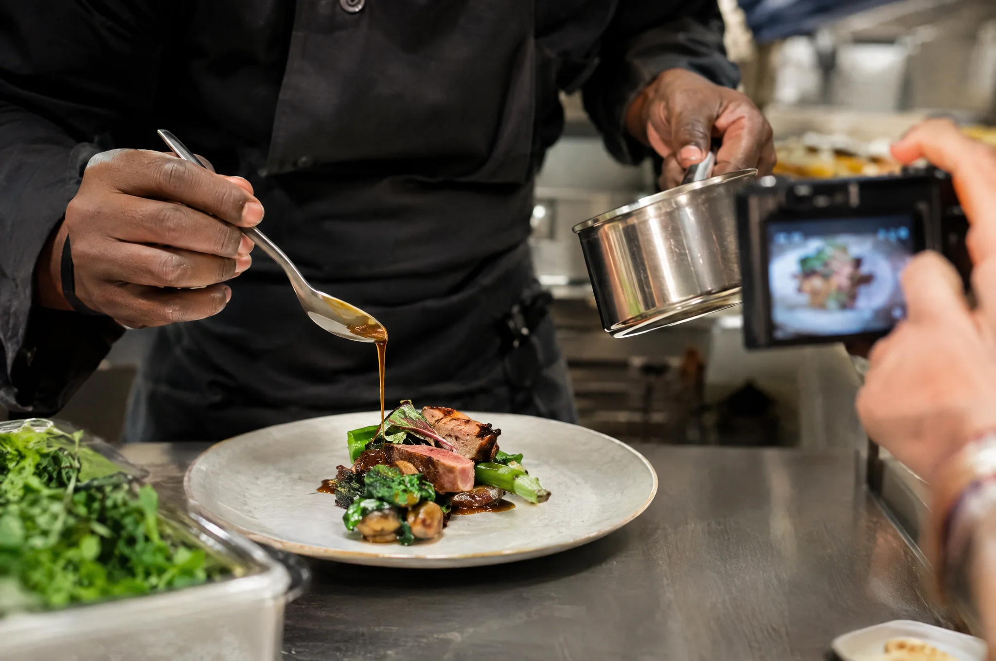 The chef finishing a plate with a spoon of sauce while a guest photographs it
