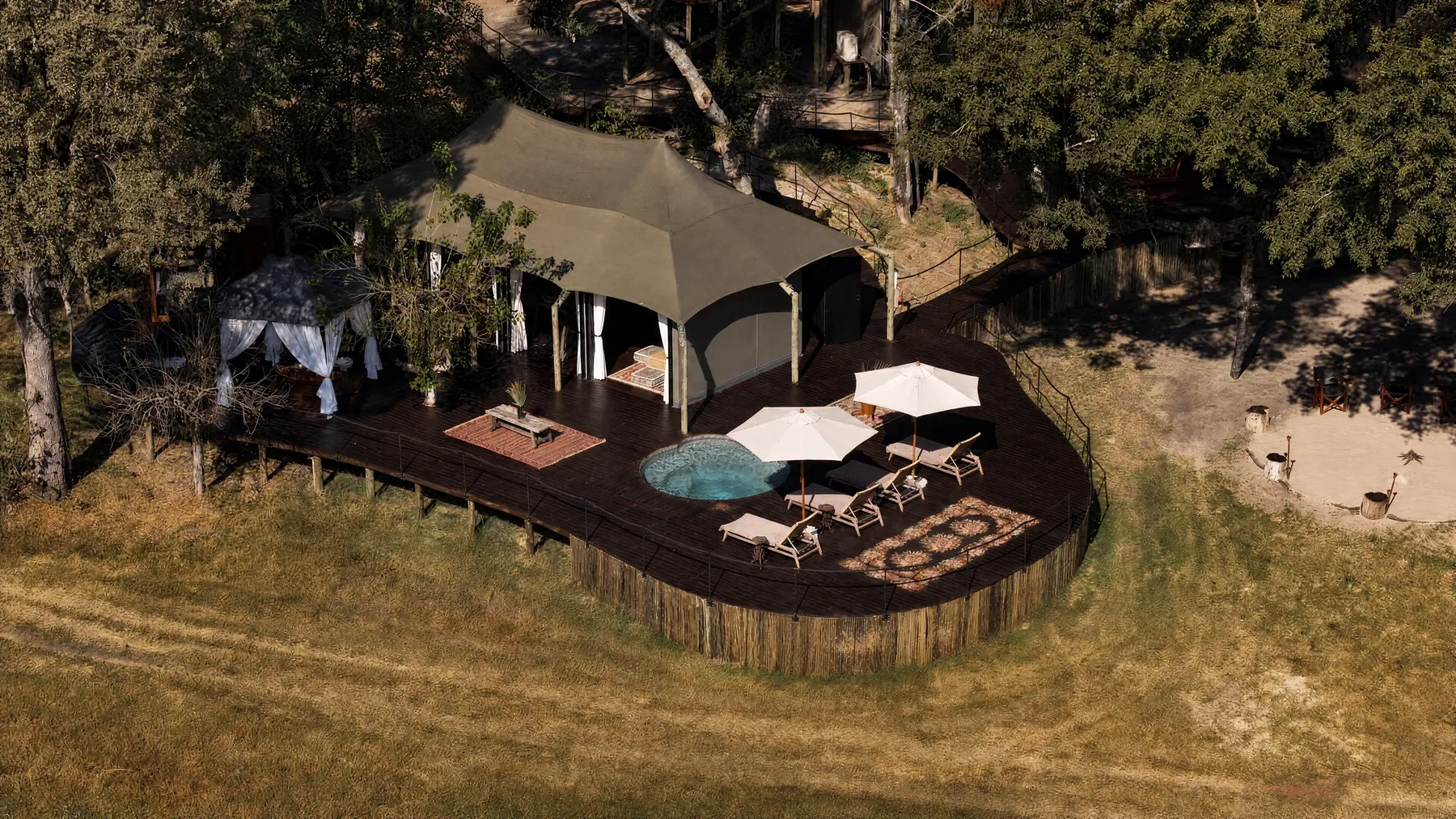 A villa's canvas house seen from the air, with its deck, a round pool and parasols, a draped pavilion beside it and a fire circle in the sand